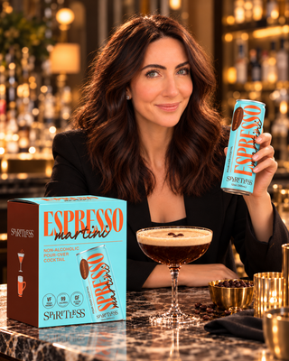 A woman in a black blazer sits at a bar holding a can of Spiritless Espresso Martini, with a matching product box and a prepared espresso martini cocktail on a marble counter in front of her.
